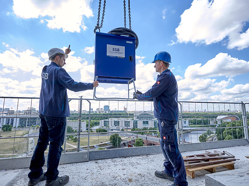2 Mitarbeiter in blauer Arbeitskleidung und mit Baustellenhelm stehen auf einem Dach einer Baustelle und positionieren Hardware von EGB