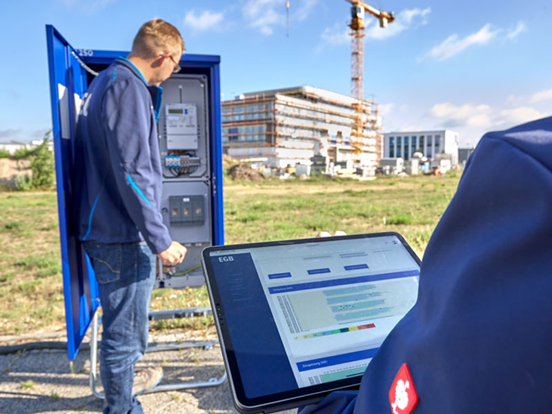 Zwei Mitarbeiter in blauen Arbeitsjacken stehen an einer Baustelle und werten die Daten des Energieverbrauchs aus.