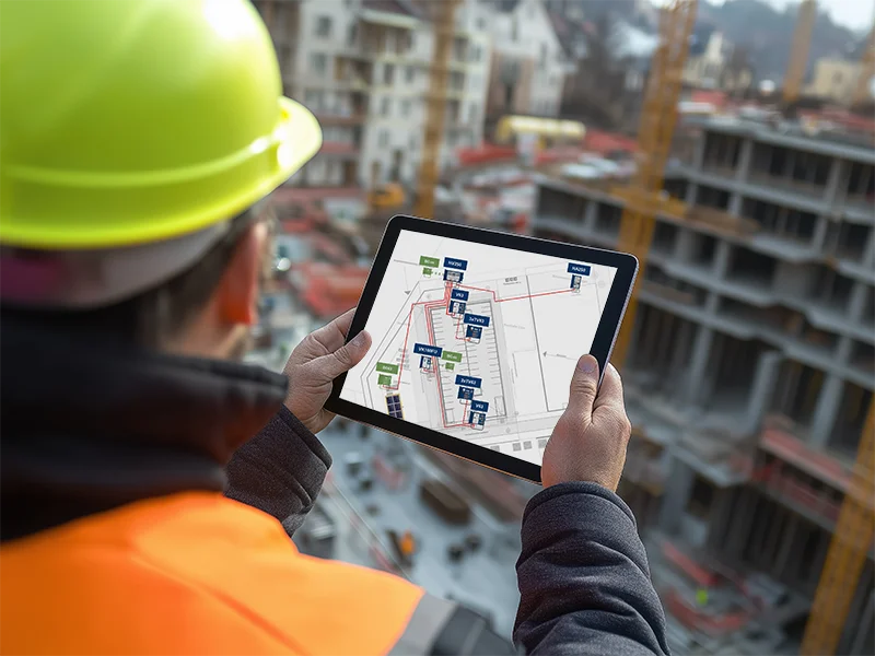Ein Mann steht mit dem Tablet in der Hand auf einer Baustelle und überblickt das Areal sowie die Stromauswertung auf dem Tablet